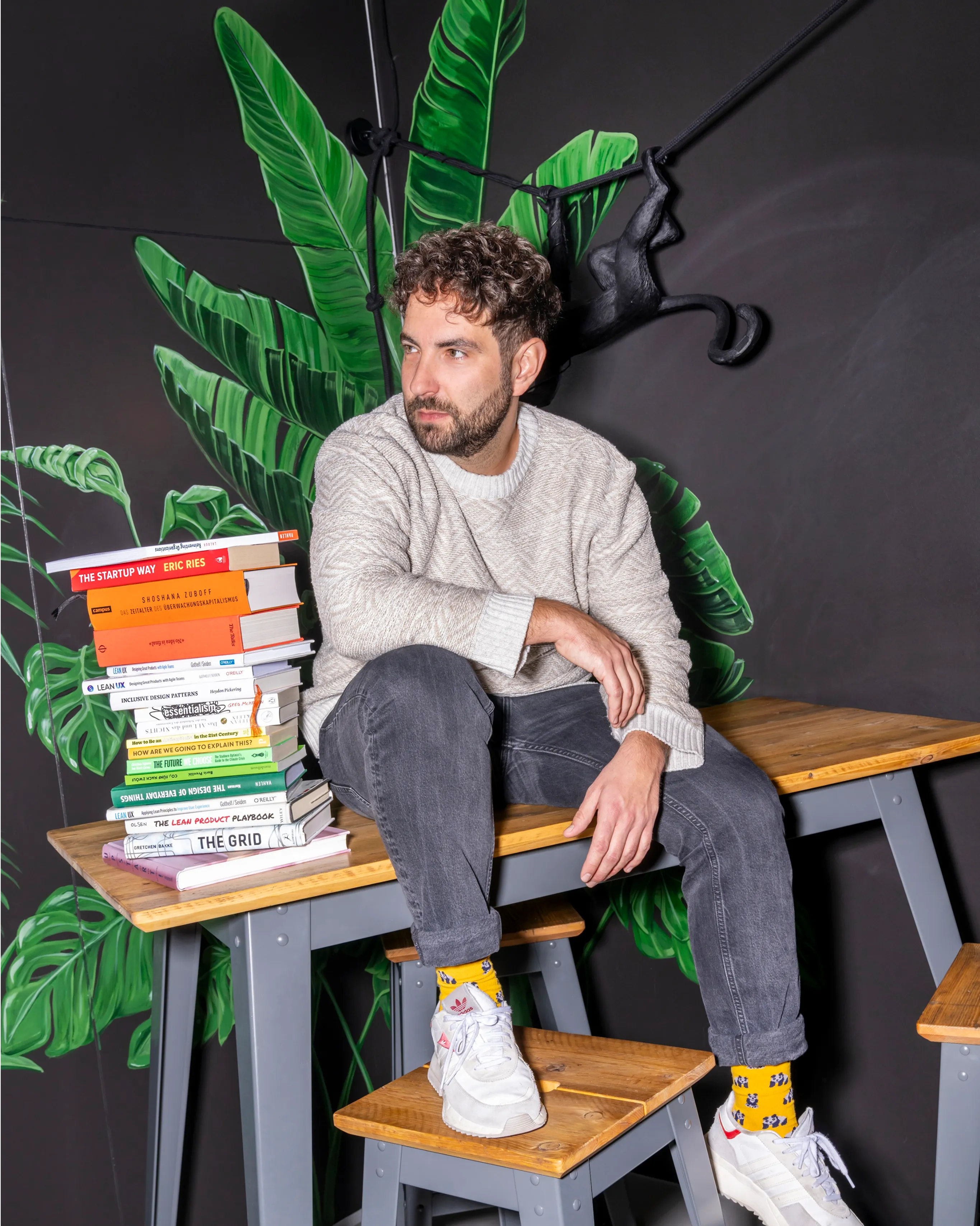Ein Foto von einem Mann mit dunklen Locken der auf einem Tisch sitzt und neben ihm ist ein Stapel bunter Bücher.