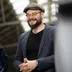 Ein Mann mit Bart, runder Brille und grauer Schiebermütze steht lächelnd in einem Innenraum mit Glasdach. Er trägt ein schwarzes T-Shirt unter einem blauen Sakko und hat die Hände vor dem Körper verschränkt. Im Hintergrund sind weitere Personen und eine Metallkonstruktion zu sehen.
